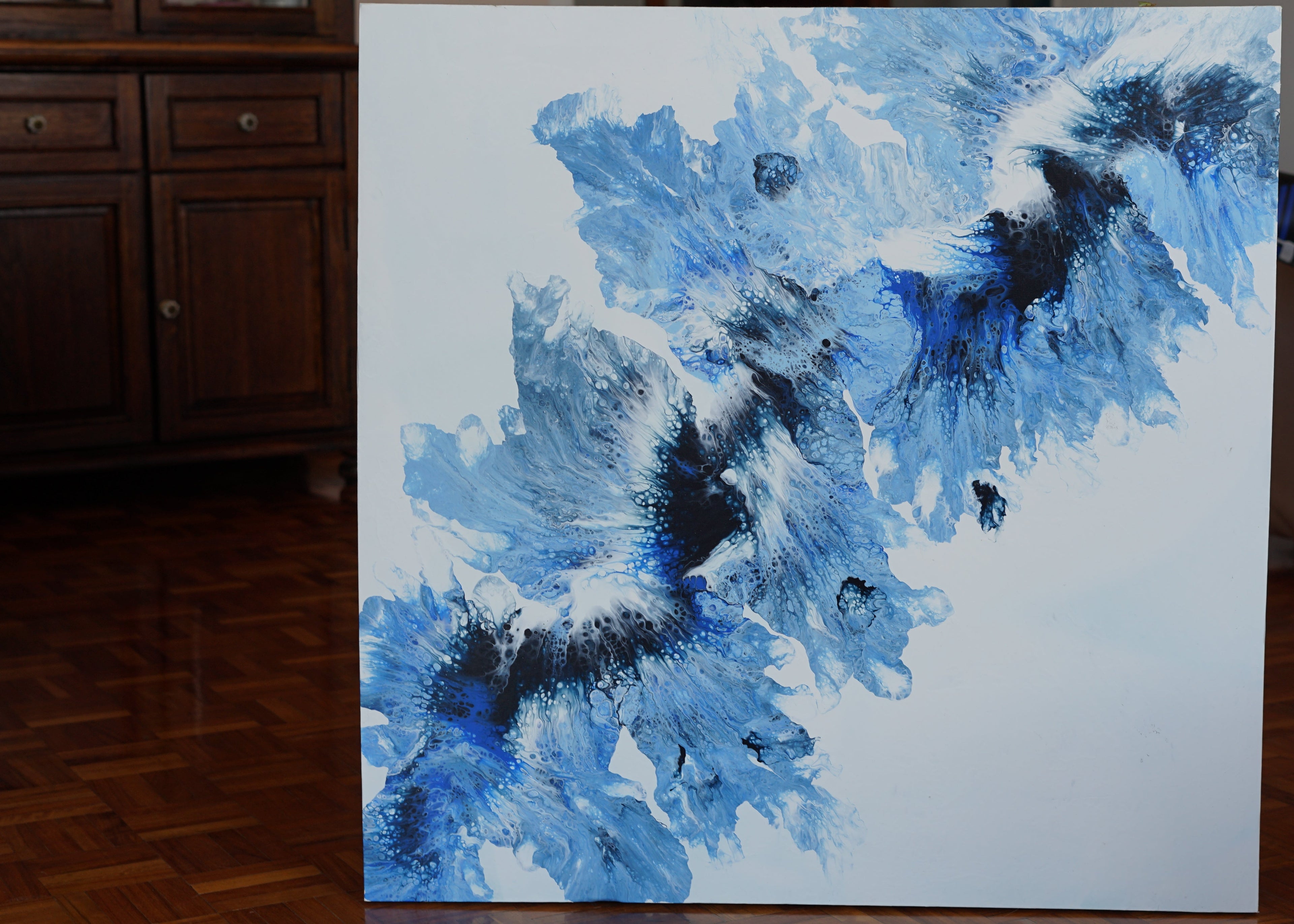 Abstract blue and black painting on a white canvas in a room with wooden flooring and furniture.
