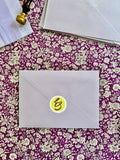 A light lilac envelope with a round yellow and black monogram sticker sits on a purple and gold floral patterned fabric surface, with stacked envelopes and a wax seal stamp visible in the background.